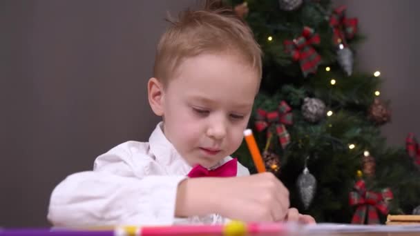Bom menino pinta cartão de saudação para os pais para o Natal com lápis de cor. Criança escreve carta com desejos ao Papai Noel, árvore de Natal decorada no fundo — Vídeo de Stock
