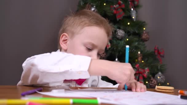 Menino em roupa festiva com gravata borboleta senta-se pensativo com caneta na mão e escreve carta para o Papai Noel sobre presentes, ou faz lição de casa na escola durante as férias de Natal — Vídeo de Stock