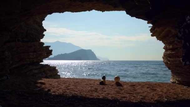Dachshund в футболке сидит у входа в грот на песчаном пляже после игры с мячом, и любуется красивым морским пейзажем, редким видом изнутри пещеры. Тогда собака убегает. — стоковое видео
