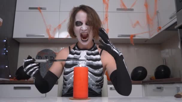 Młoda kobieta z makijażem czarownicy Halloween i kostiumem, z brutalnym wyrazem twarzy, wykonując okrągły ruch rąk powyżej grzania noża na płomieniu świecy. Procedura czarnoksięstwa, pojęcie humoru — Wideo stockowe