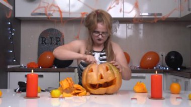 Cadılar Bayramı cadı kostümü giymiş genç ve komik bir kadın ve sofistike bir şekilde süslenmiş mutfakta oturmuş, balkabağını güçlü bir şekilde içine sürterek balkabağı yapıyor.