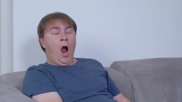Hombre cansado bosteza sentado en cómodo sofá gris en la habitación — Vídeos de Stock