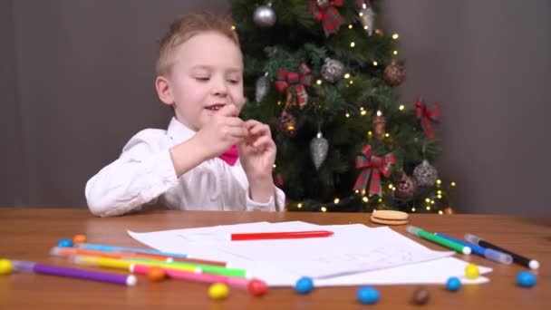 Little boy eats candy at table near Christmas tree at home — Stock Video