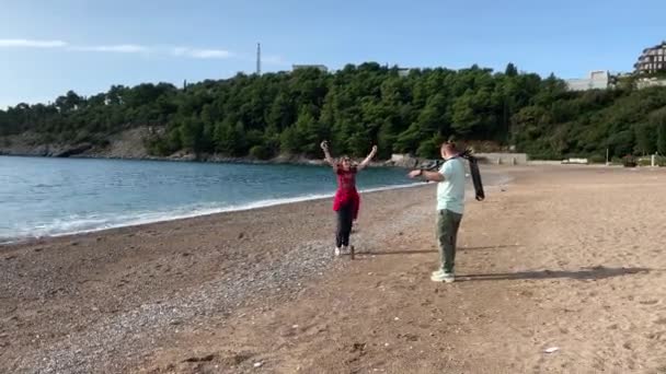 Pareja joven estaba caminando en la playa y montar scooter, se detuvieron para que ese tipo tomó una foto de la mujer saltando con alegría contra el fondo del mar para su blog — Vídeo de stock