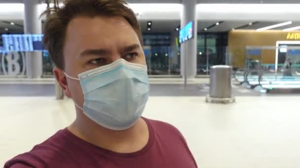 Молодий чоловік в захисній медичній масці на обличчі йде по аеропорту і з ентузіазмом розмовляє, жестикулюючи руками і роблячи жест з головою, вказуючи напрямок — стокове відео
