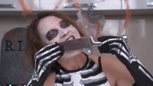 Mujer loca joven con maquillaje espeluznante y disfraz de esqueleto maníaco lame cuchillo afilado. Actriz escenificada para amigos en fiesta de Halloween — Vídeo de stock