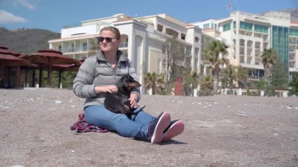 Una joven con gafas de sol se sentó sobre una manta para descansar al sol mientras caminaba con su mascota. propietaria femenina sostiene lindo perro dachshund obediente en sus brazos — Vídeos de Stock