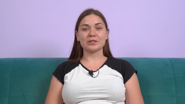 Pozytywne blogerka młoda kobieta z mikrofonem na szyi koszulki rozmowy uśmiecha się do kamery na miękkiej zielonej kanapie w pobliżu fioletowej ściany zbliżenie. Nagrywanie kursu online — Wideo stockowe