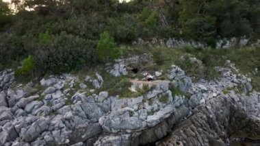 Genç aktif adam ağaçlarla çevrili bir platformda oturuyor. İnsansız hava aracı kayaları yıkarken deniz manzarasını gözler önüne seriyor. Gezgin blogu için ulaşılması zor güzel yerleri keşfeder.