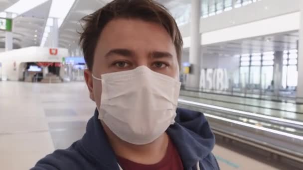 Ung man i medicinsk mask som skyddar honom från virus och förorenad miljö går genom den stora hallen på flygplatsen, och talar entusiastiskt med förvånad blick, framifrån — Stockvideo