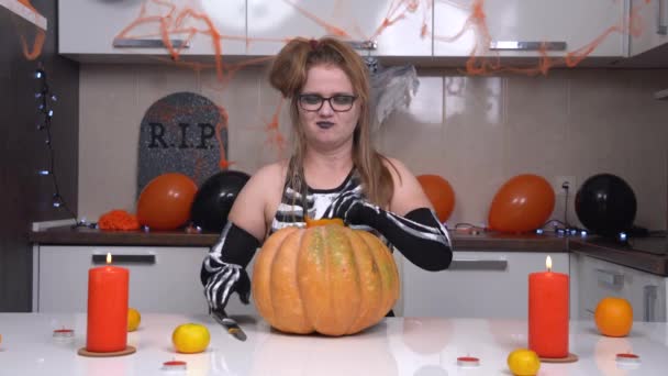 Jovem fêmea engraçada em traje de bruxa de Halloween e maquiagem sentada na mesa na cozinha decorada para férias, em cores preto e laranja, corta uma peça de abóbora grande, sorrindo ameaçadoramente — Vídeo de Stock