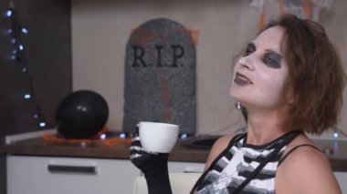 Ürkütücü makyajlı ve iskelet kostümlü genç bir kadın Cadılar Bayramı partisinde içtiği bardaktan ferahlatıcı, rahatlatıcı ya da sıcak bir içecek, dekore edilmiş bir daire.