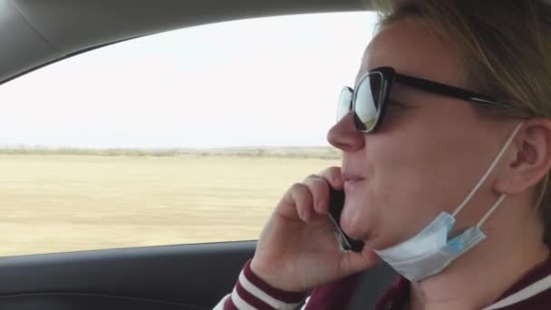 Jonge blonde vrouw met een zonnebril beëindigt een gesprek op mobiele telefoon, knikt bevestigend met haar hoofd en wijst het gesprek af, draagt een beschermend medisch masker op haar gezicht, beweegt zich in de auto — Stockvideo