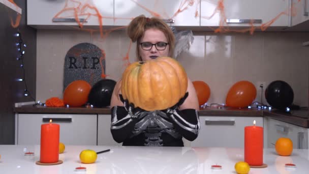 Joven hembra divertida en disfraz de bruja de Halloween y maquillar la celebración de la calabaza grande en la mesa en la cocina decorada para las vacaciones, en colores negro y naranja, con globos, guirnaldas, velas y frutas — Vídeos de Stock