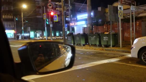 Espejo lateral de un coche, de pie en una intersección en el semáforo rojo, vista desde el interior del vehículo a través de la ventana. Viaje por la ciudad nocturna. Infraestructura urbana — Vídeo de stock