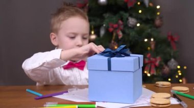 Beyaz gömlekli güzel bir çocuk ve pembe papyonlu bir kutu açar. Noel hediyesiyle birlikte Noel Baba 'ya bir dilek mektubu yazar. Arka planda süslenmiş Noel ağacı.