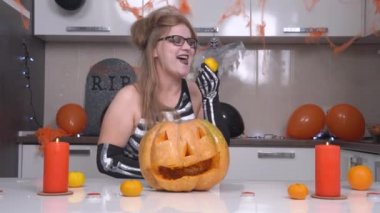 İskelet kostümlü çılgın genç kadın mandalinayı kabuğuyla yer ve ısırır. Daire, Cadılar Bayramı partisi için balkabağı Jack-o-Lantern, tüyler ürpertici örümcek ağları, mumlar ve balonlarla süslenmiş.