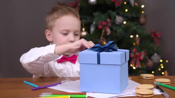 Precioso niño en camisa blanca y pajarita rosa abre caja con regalo de Navidad recibido sobre el que escribió carta con deseos a Santa, árbol de Navidad decorado en el fondo — Vídeo de stock