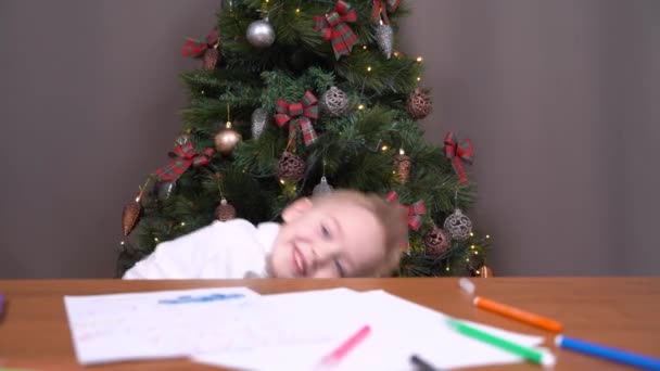 Child in festive outfit jumps out from under table showing tricks like the jack-in-the-box. Bored boy was tired of drawing and writing letter with wishes to Santa, and found new entertainment — Stock Video