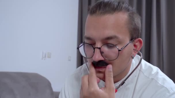 Young doctor in uniform and glasses checks health of patient using stethoscope, then rubs his thick mustache with hand in confusion. Bad news about incurable disease — Stock Video