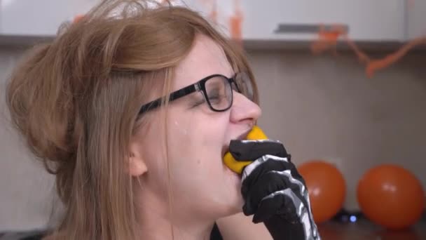 Verrückte blonde junge Frau mit seltsamem Make-up und Skeletthandschuhen beißt und isst Mandarine direkt mit Schale, aus nächster Nähe. Sie lächelt gruselig, als der Fruchtsaft über ihr Gesicht tropft — Stockvideo