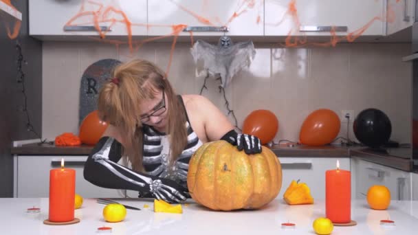 Blonde woman in creepy skeleton costume and with weird hairstyle carves face on pumpkin to make jack lantern for Halloween party. Apartment is decorated with holiday themed details — Stock Video