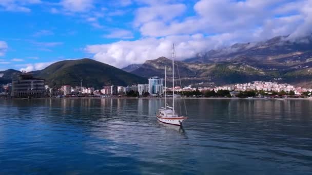 Drone lata wokół przyjemności lub łodzi rybackiej z opuszczonymi żaglami, pływające w zatoce. Europejskie miasto Budva u podnóża gór okrytych chmurami, Czarnogóra — Wideo stockowe