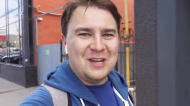 Modern kablosuz kulaklıklı genç adam sokakta yürüyor ve el kol hareketi yaparken duygusal konuşuyor. Video blogcusu gülümsüyor ve komik bir hikaye anlatıyor