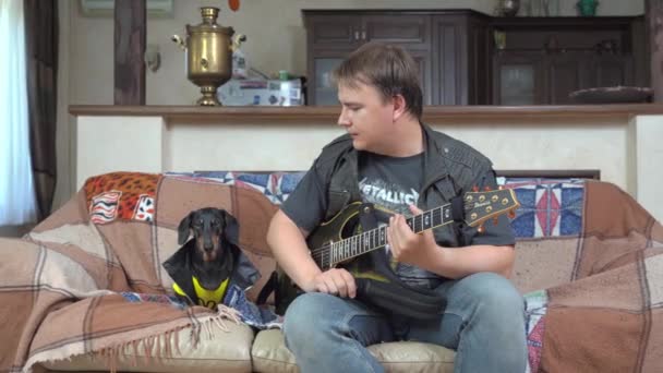 5月25日、ロシア、ロストフ:エレキギターを持つ若い男は、彼のロックパフォーマンスやコンサートのリハーサルを見るために革のジャケットで面白いダックスフンド犬を呼び出します。趣味・娯楽の概念 — ストック動画