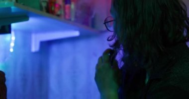 Gözlüklü adam yarı karanlık odada elektrikli sigara içiyor.