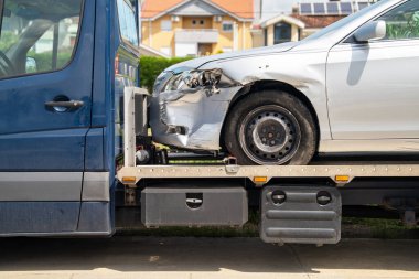 Çarpışma hasarı olan gümüş bir arabanın ön sol tarafının daha yakın görüntüsü ve yerleşim yerinin önündeki çekici platformuna bağlanmış bir tampon.