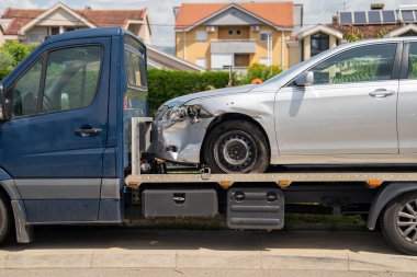 Önü görünen bir çarpışma hasarı olan gümüş bir araba, arka planda evleri olan bir banliyö caddesinde park halindeki bir çekici kamyonunda emniyete alındı.