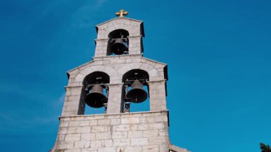 Açık mavi bir gökyüzünün altında, üzerinde güneşte parıldayan küçük altın bir haç bulunan üç kemerli ve dört çanı olan uzun bir Ortodoks çan kulesi yükseliyor..