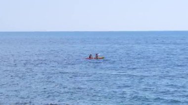 İki birey, berrak bir gökyüzünün altında, sakin bir mavi okyanusun ortasında küçük bir kanoyla kürek çeker. Açık suların engin genişliğinde uzak görünür.