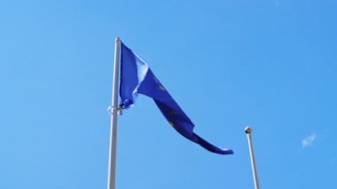 Açık mavi gökyüzüne karşı rüzgarda dalgalanan sarı yıldızlı bir Avrupa Birliği bayrağı, açık havada uzun beyaz bir bayrak direğine monte edilmiştir.