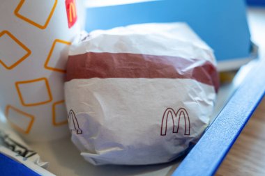 Belgrad, Sırbistan: 3 Temmuz 2024: Kağıda sarılmış bir McDonalds burgeri, tek kullanımlık bir içecek bardağının yanında, masada fast food menüsü