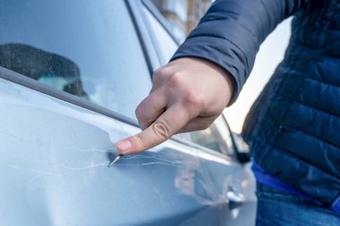 Gümüş renkli bir arabanın kapısındaki boyayı çizmek için metal bir anahtar kullanan bir kişinin yakın plan görüntüsü. Vandalizm eyleminde uzun çizgiler oluşturur. Ceket kolu görünür ve araç yüzeyinde yansımalar olur.