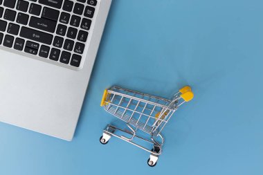 Düz dizüstü bilgisayar klavyesi ve mavi arka planda minyatür alışveriş arabası. Çevrimiçi alışveriş, e-ticaret, dijital perakende satış ve bir bilgisayardan malzeme sipariş etme kavramı.