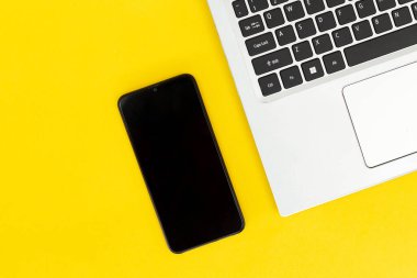 Düz dizüstü bilgisayar, parlak sarı arka planda boş ekranlı siyah akıllı telefon. Modern ofis veya dijital uzaktan kumanda kurulumu kavramı.