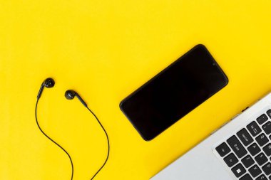 Düz dizüstü bilgisayar, boş ekranlı siyah akıllı telefon ve parlak sarı arka planda kabloyla kulaklık. Dijital çalışma veya müzik akışı kurulumu kavramı