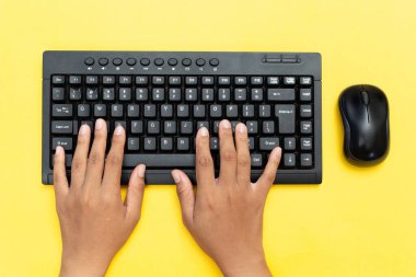 Siyah klavyede yazı yazan ellerin üst görüntüsü ve canlı sarı arka planda bir fare kullanımı. Veri girişi, ofis çalışması veya uzaktan üretkenlik kavramı