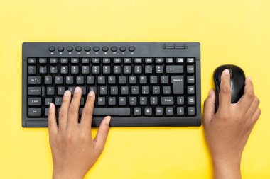 Siyah klavyede yazı yazan ellerin üst görüntüsü ve canlı sarı arka planda bir fare kullanımı. Veri girişi, ofis çalışması veya uzaktan üretkenlik kavramı