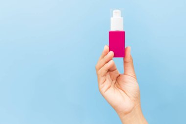 Temiz açık mavi arka planda pembe beyaz bir kozmetik pompa şişesini tutan el. Minimalist cilt ürünü kopyalama alanı ile maketi