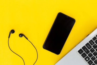 Düz dizüstü bilgisayar, boş ekranlı siyah akıllı telefon ve parlak sarı arka planda kabloyla kulaklık. Dijital çalışma veya müzik akışı kurulumu kavramı