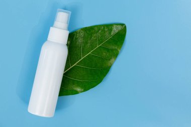 Taze yeşil bir yaprağın üzerinde beyaz plastik sprey şişesi, doğal cilt bakımını ve organik güzellik ürünlerini temsil eden saf mavi bir arka planı var..