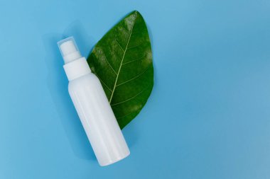 Taze yeşil bir yaprağın üzerinde beyaz plastik sprey şişesi, doğal cilt bakımını ve organik güzellik ürünlerini temsil eden saf mavi bir arka planı var..