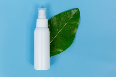 Taze yeşil bir yaprağın üzerinde beyaz plastik sprey şişesi, doğal cilt bakımını ve organik güzellik ürünlerini temsil eden saf mavi bir arka planı var..