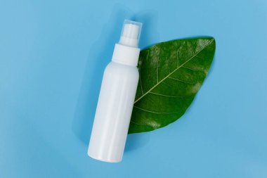 Taze yeşil bir yaprağın üzerinde beyaz plastik sprey şişesi, doğal cilt bakımını ve organik güzellik ürünlerini temsil eden saf mavi bir arka planı var..