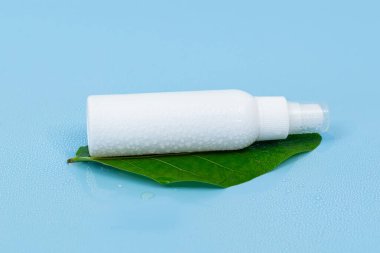 Taze yeşil bir yaprağın üzerinde beyaz plastik sprey şişesi, doğal cilt bakımını ve organik güzellik ürünlerini temsil eden saf mavi bir arka planı var..