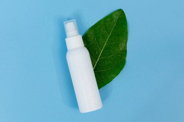 Taze yeşil bir yaprağın üzerinde beyaz plastik sprey şişesi, doğal cilt bakımını ve organik güzellik ürünlerini temsil eden saf mavi bir arka planı var..